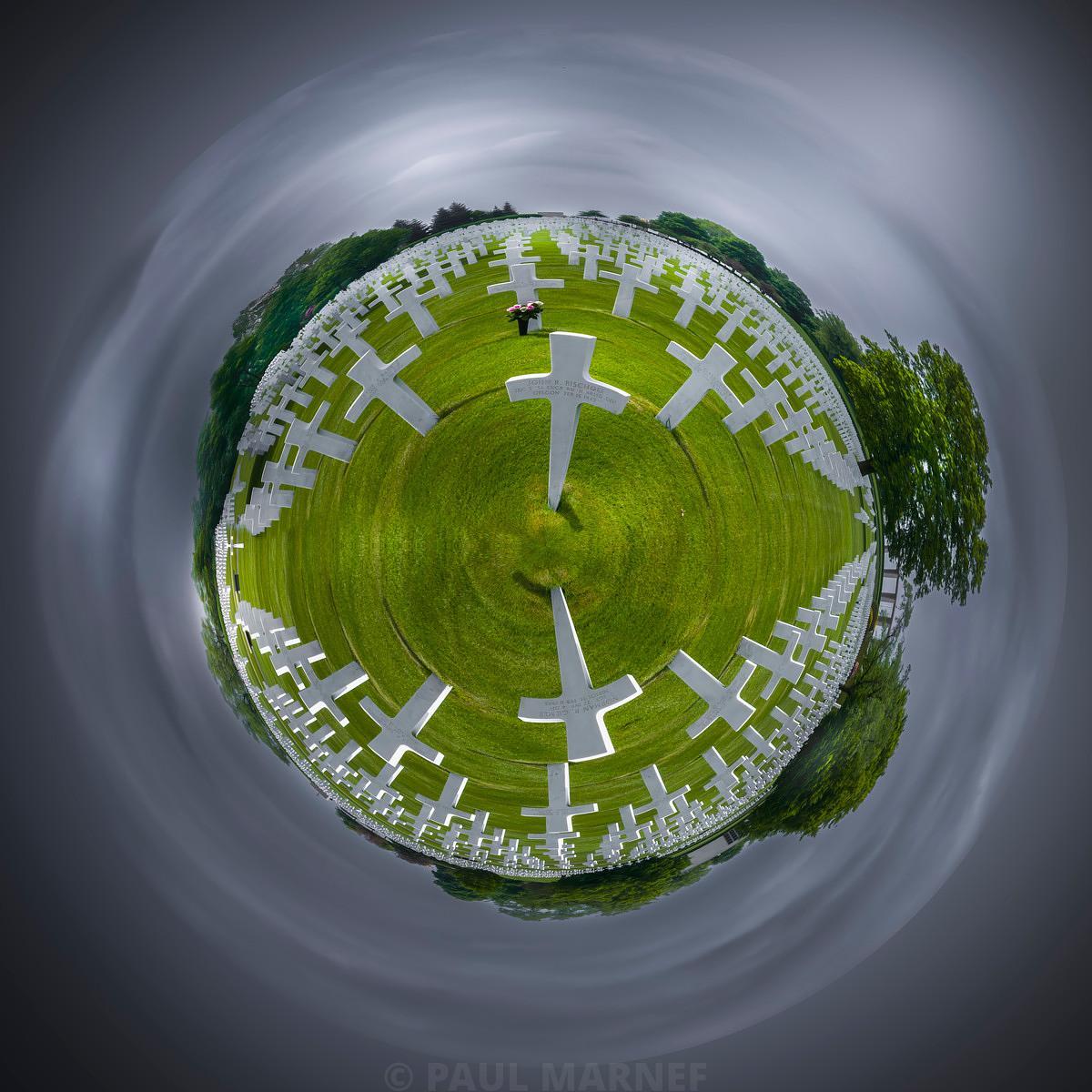 A spherical aerial view of a vast open-air cemetery, where dozens of white crosses are scattered across a green lawn beneath a grey, cloudy sky.