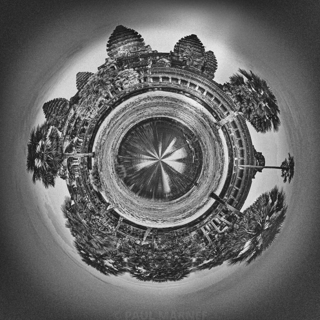 A distorted black-and-white panoramic view of the stone towers of Angkor Wat, surrounded by palm trees and tropical foliage.