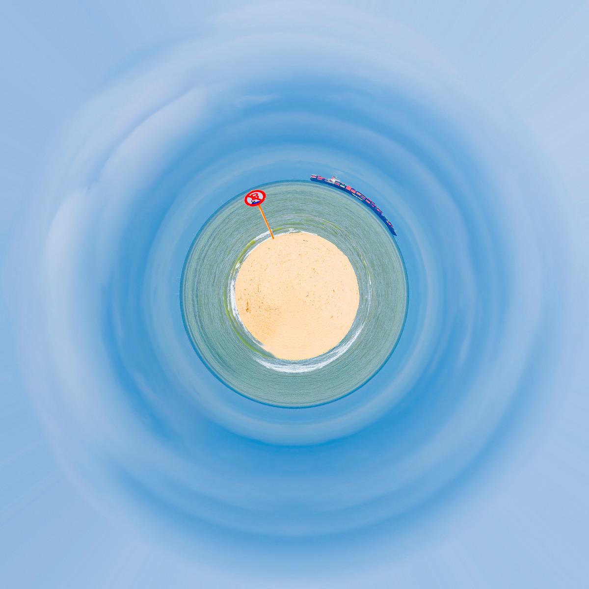 Vue plongeante sur une plage d'île entourée de lagon turquoise, un cargo sur l'eau et un pylône rouge isolé sur le sable.