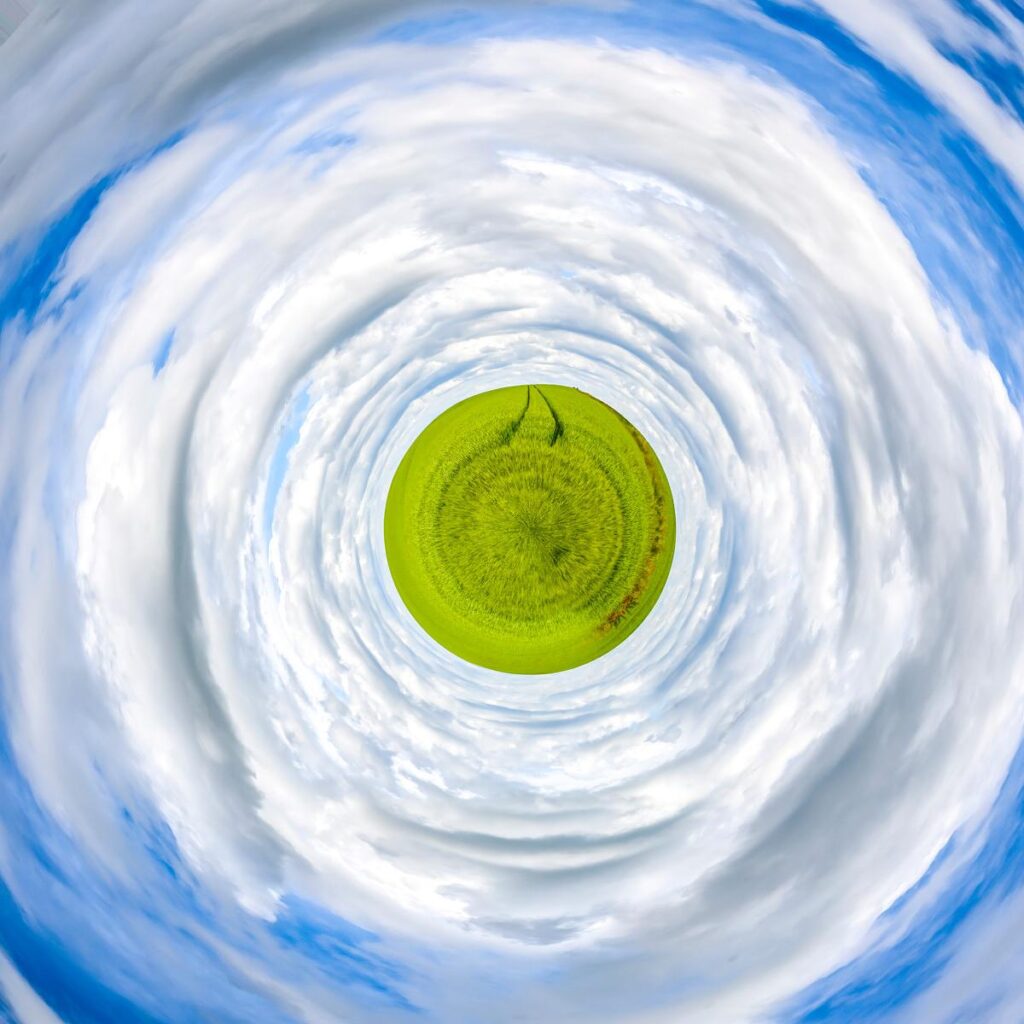 A spiral vortex effect formed by white clouds and a blue sky, drawing the eye towards a central patch of green meadow.