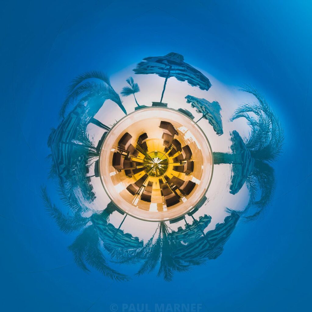 A circular reflection of parasols and palm trees at the edge of a turquoise-blue swimming pool.