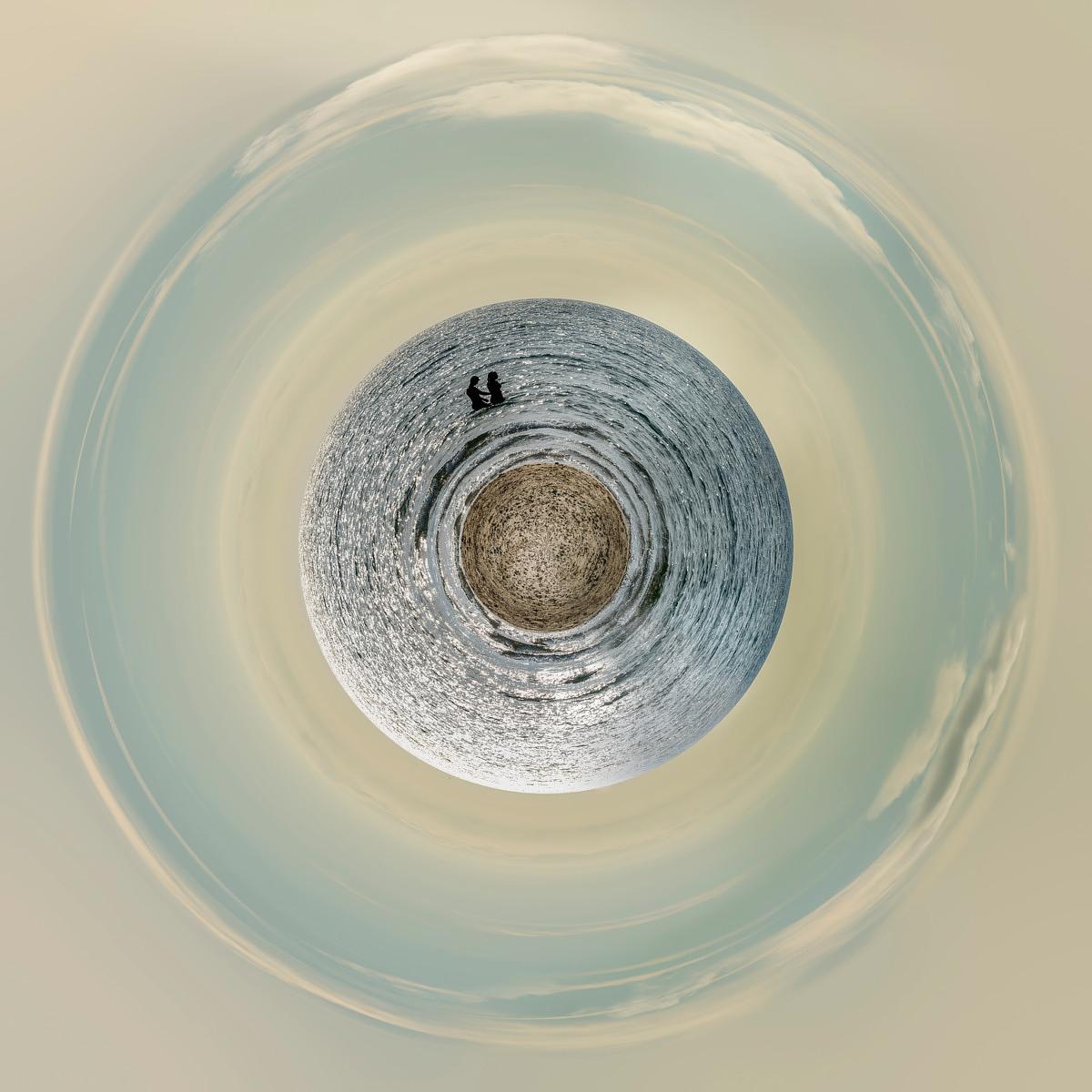 Silhouette de deux personnes sur une masse d'eau sphérique, enveloppée de vagues concentriques et d'un ciel pâle.