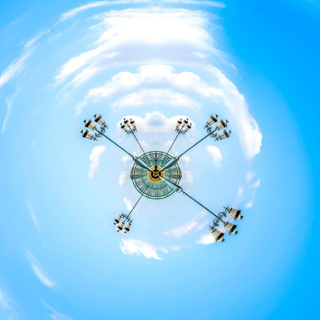A bird’s-eye view of the top of a wheel adorned with white street lamps, set against a bright blue sky dotted with white clouds