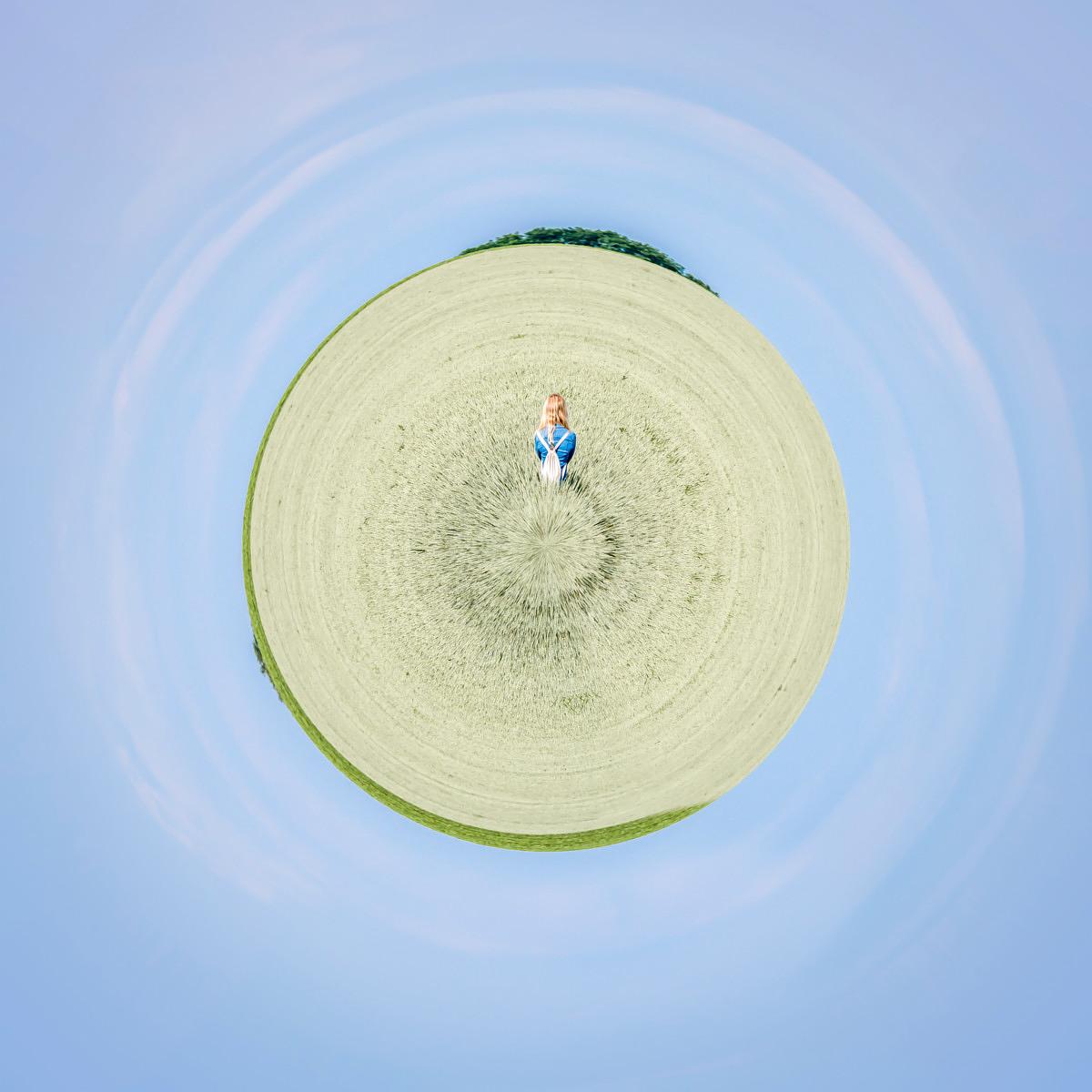 Une femme rousse debout au centre d'un champ de céréales en forme de sphère flottant dans un ciel bleu clair.