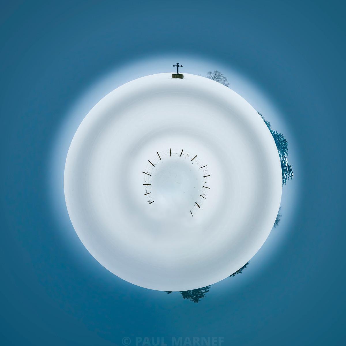 Un vaste ciel bleu enveloppe un disque neigeux surmonté d'une croix, entouré de silhouettes d'arbres et de petits traits noirs.