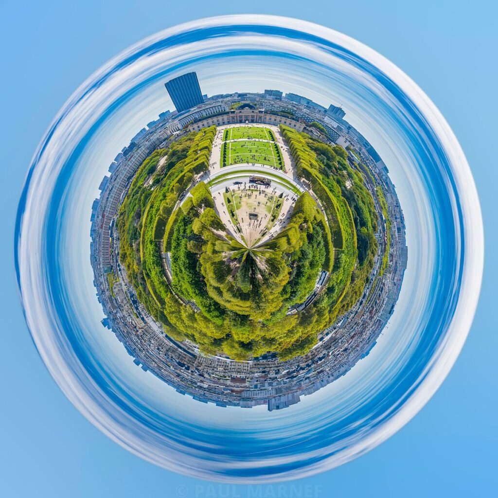 Vue aérienne circulée où un parc verdoyant et des allées blanchâtres forment un disque central entouré d'un cortège de toits urbains et d'un grand ciel bleu partiellement voilé par des nuages étirés.