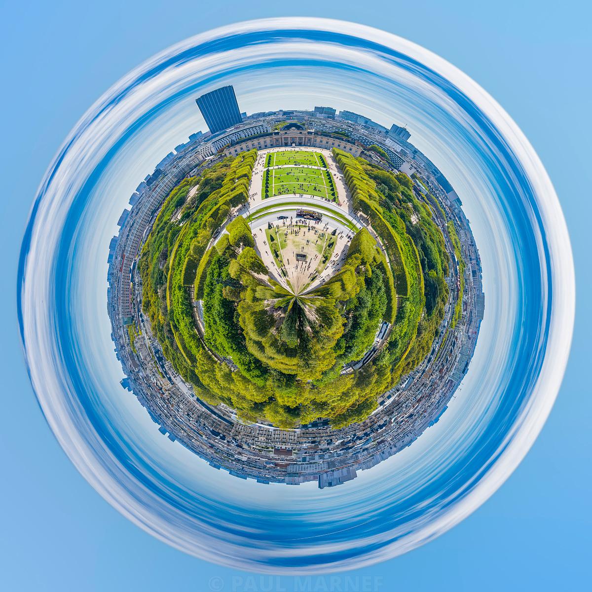 Vue aérienne circulée où un parc verdoyant et des allées blanchâtres forment un disque central entouré d'un cortège de toits urbains et d'un grand ciel bleu partiellement voilé par des nuages étirés.