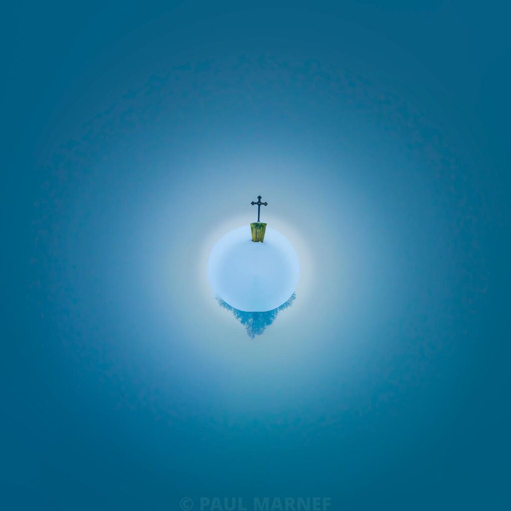 A vertical bird’s-eye view of the green spire of a stone cross topping a bell tower, standing alone against a clear blue sky with wisps of mist.