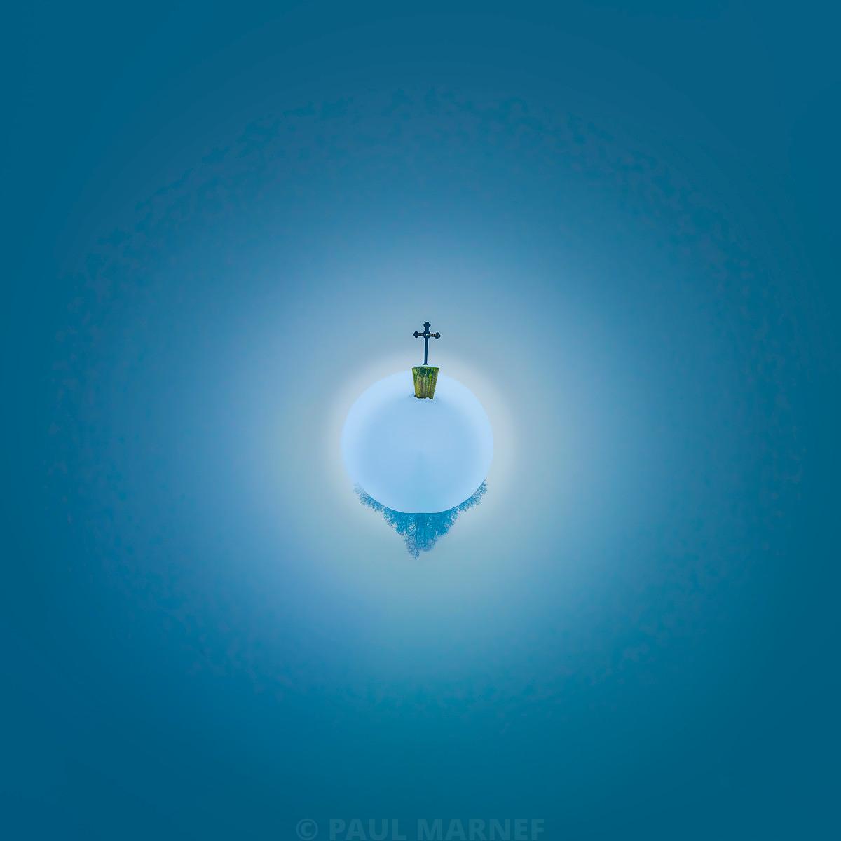 Une vue verticale plongeante sur le pinacle vert d'une croix de pierre surmontant un clocher, isolé dans un ciel bleu clair aux légères nappes de brume.