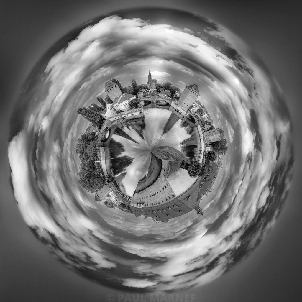 A village surrounded by gardens forming a circle against a swirling sky, with clouds creating a spiral effect in black and white.