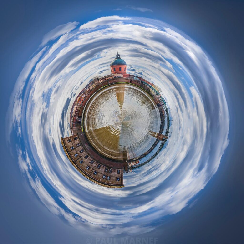 Vue 360 degrés d'une place avec une fontaine et une architecture aux dômes roses, le ciel et ses nuages s'enroulent en spirale.