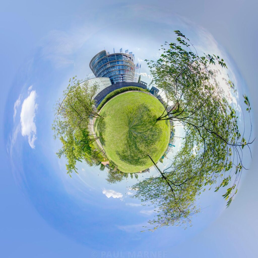 A panoramic view showing a curved, glass-fronted building in the centre of the sky, surrounded by a green lawn and bare branches.