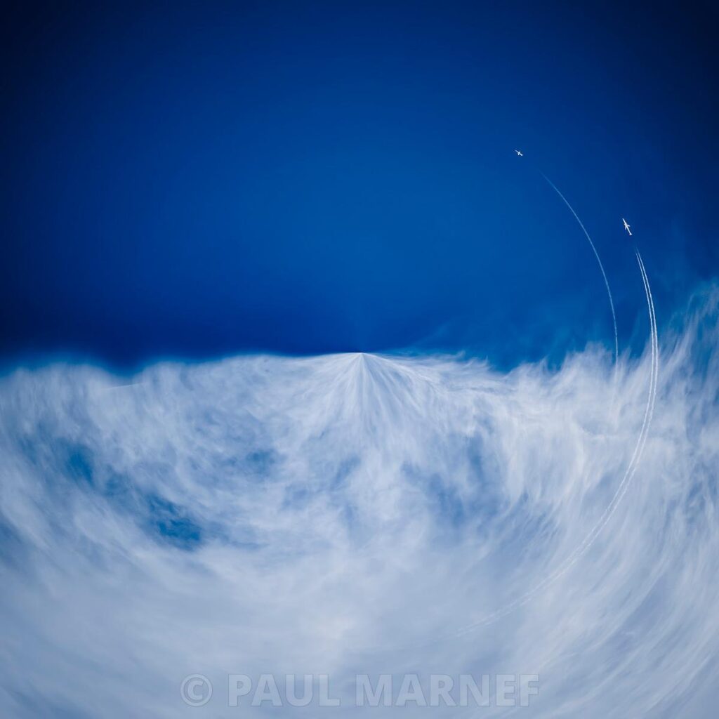 Two aeroplanes are leaving white, arched contrails across a blue sky, flying above thin, dense clouds.