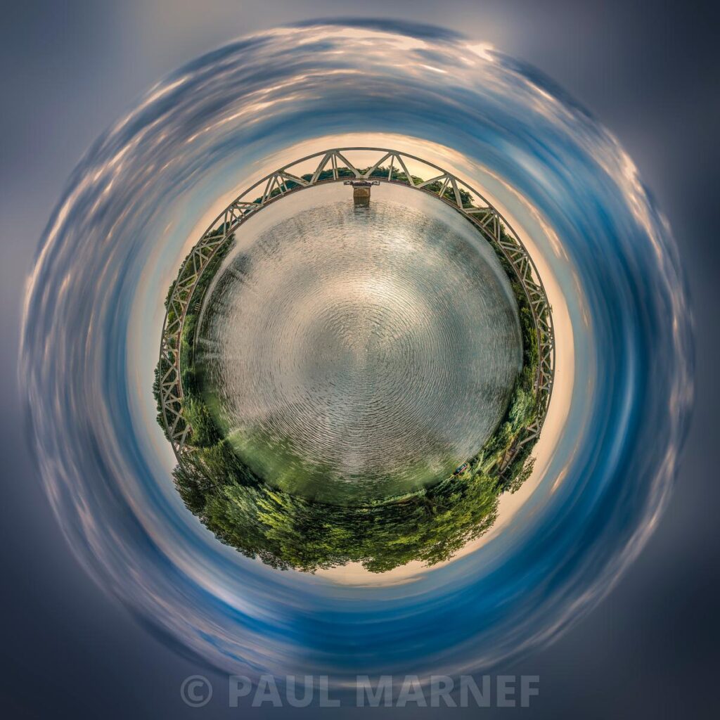 A vertical view of a circular concrete pool surrounded by a metal structure and dark green vegetation, set against a backdrop of a blue sky swirling with wisps of whitish clouds.