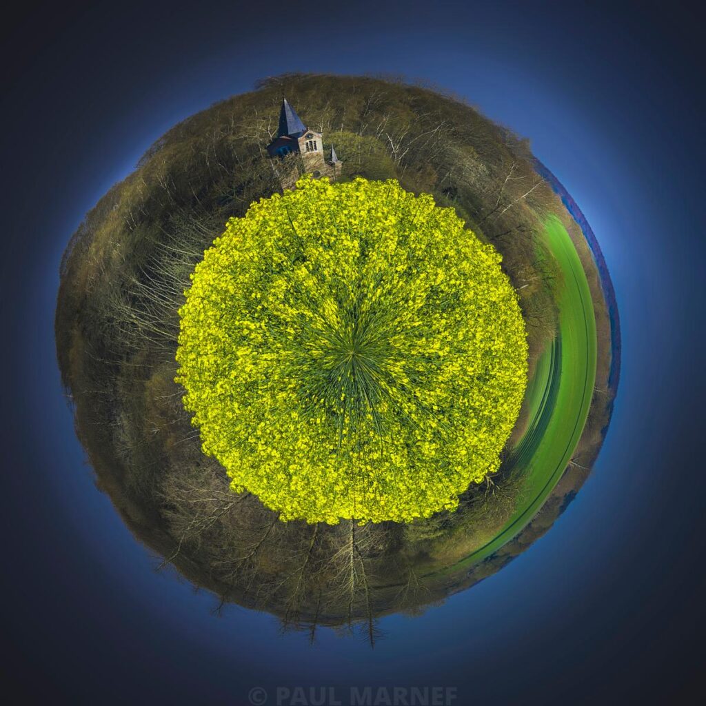Projeté sur un monde miniature, le clocher d'une église surplète un champ de colza jaune pétant encadré de forêts, sous un ciel bleu sombre.