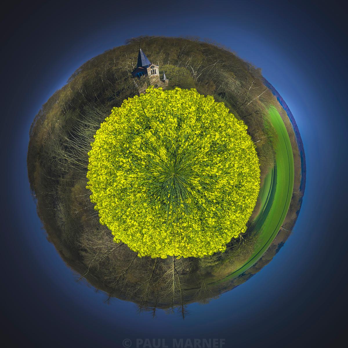 Projeté sur un monde miniature, le clocher d'une église surplète un champ de colza jaune pétant encadré de forêts, sous un ciel bleu sombre.