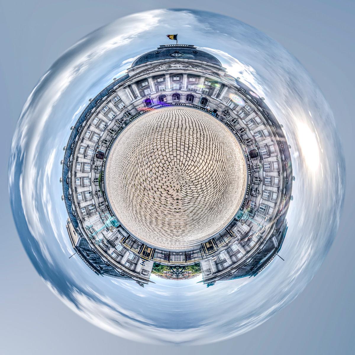 The façade of a Parisian building and a courtyard paved in a chequered pattern curve into a sphere against a backdrop of a blue-grey, cloudy sky.