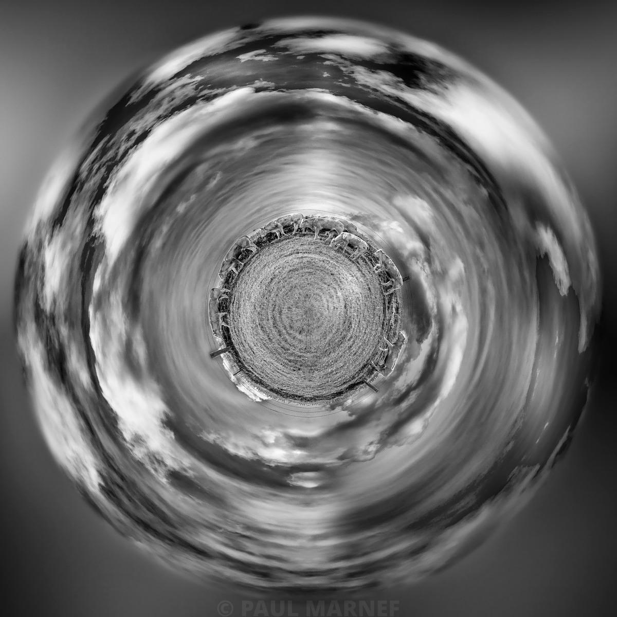 Vue circulaire noir et blanc d'une meule de foin au centre d'une plaine, sous un ciel où les nuages se déploient en spirale.