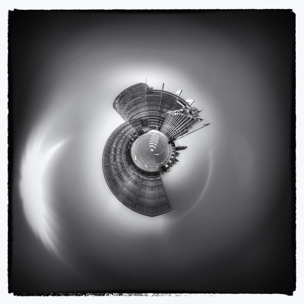 Édifice en courbe et dôme central isolés dans un ciel nuageux, capturé en noir et blanc avec un effet optique de distorsion circulaire.