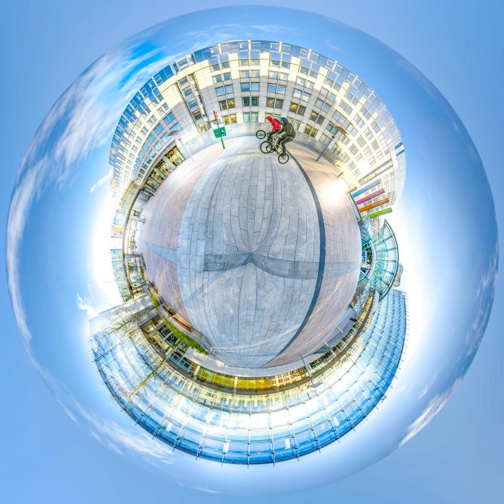 A view in the shape of a small globe showing a curved, glass-fronted building in a square, with two cyclists riding past the façades and a blue sky in the background.
