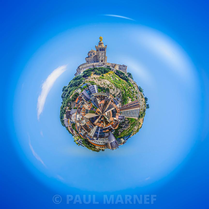 La basilique Notre-Dame de la Garde surplombe un village de toits et de verdure dans une perspective circulaire sous un ciel bleu vif.