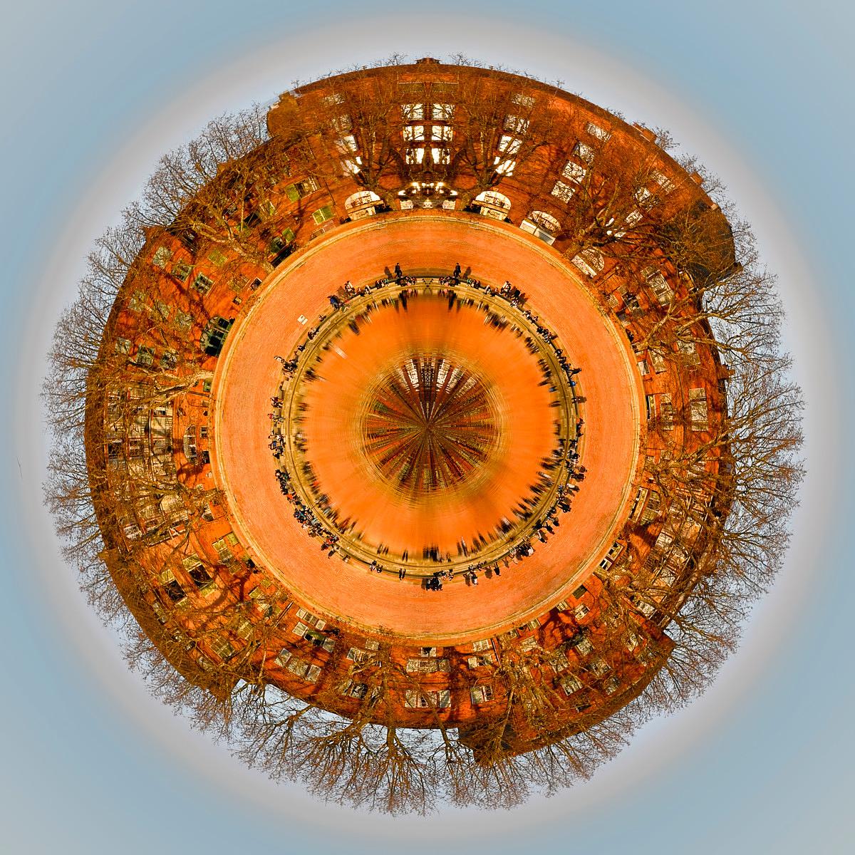 Vue en projection 360 degrés d'un parvis circulaire en pavés orangés avec des silhouettes de personnes et des immeubles aux façades orangées entourés de arbres sans feuilles sous un ciel bleu clair.