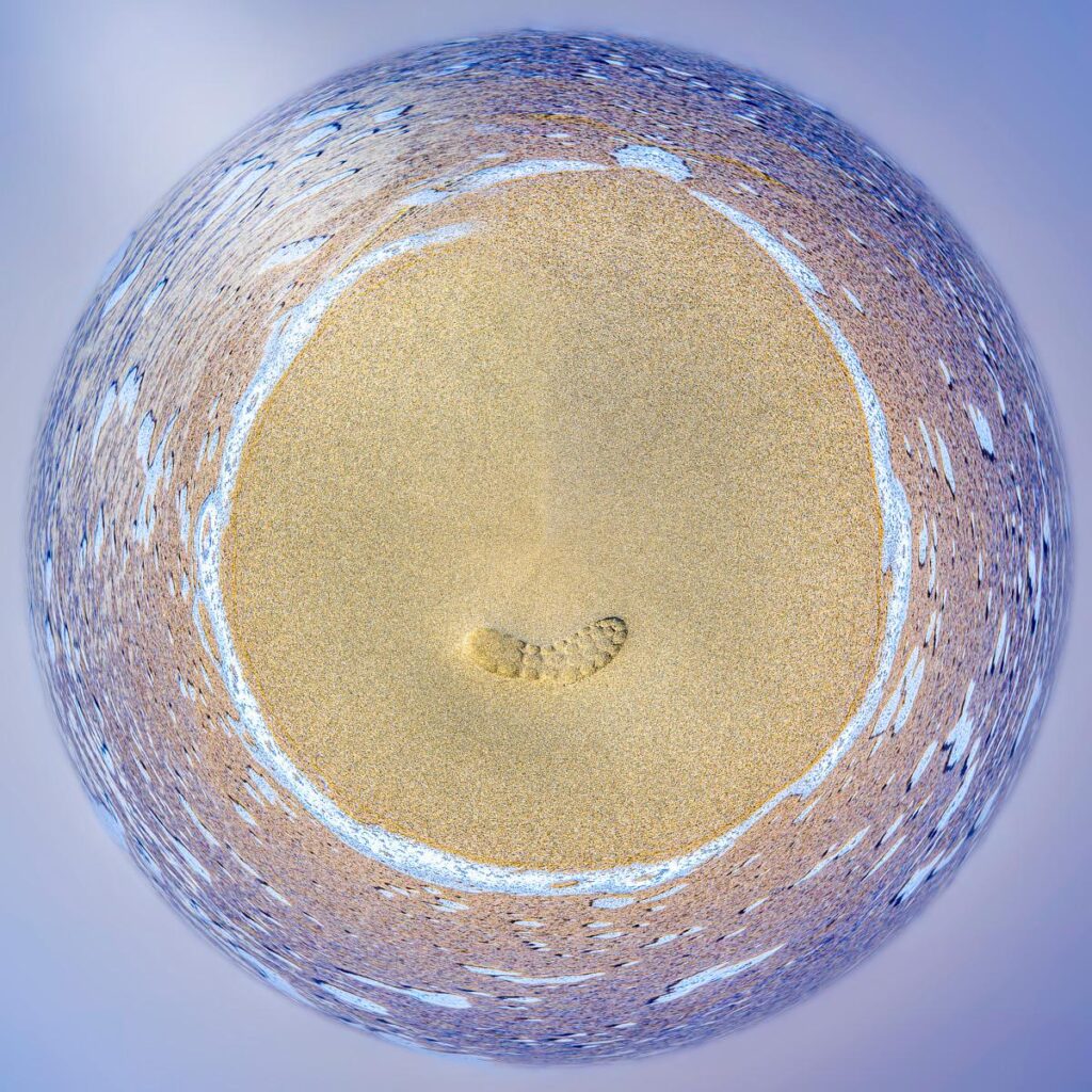 Vue panoramique circulaire d'une plage avec une empreinte dans le sable entourée d'une vague d'écume sur une mer agitée.