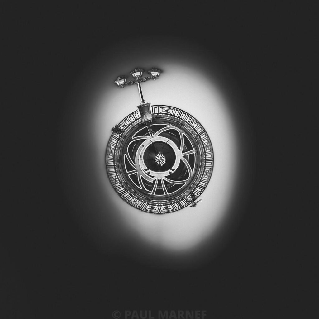 Vue d'une lucarne circulaire au-dessus de la tour Eiffel, avec sa structure en entrelacs métalliques sous un halo lumineux monochrome.