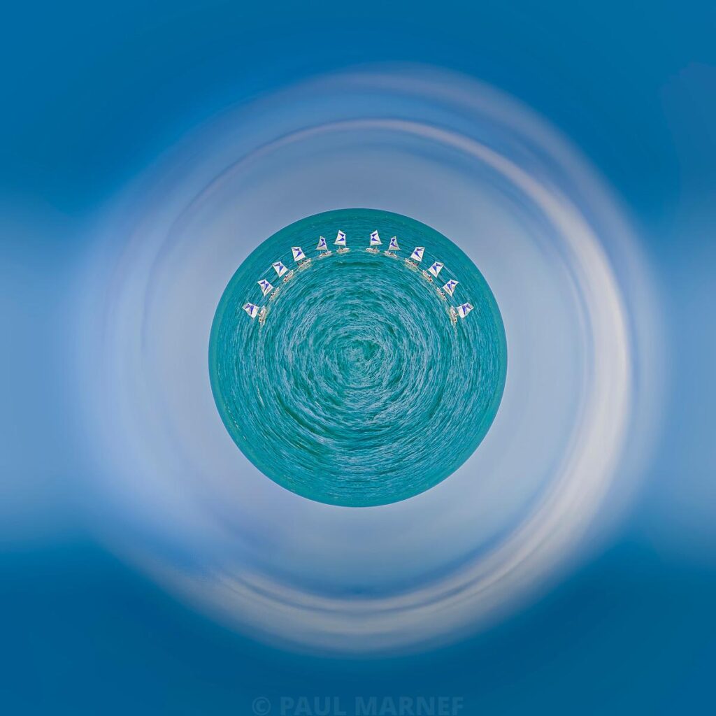 Une flottille de petits voiliers cerne un disque d'eau turquoise et tournoyante, entourée de larges flous circulaires dominés par le bleu ciel.