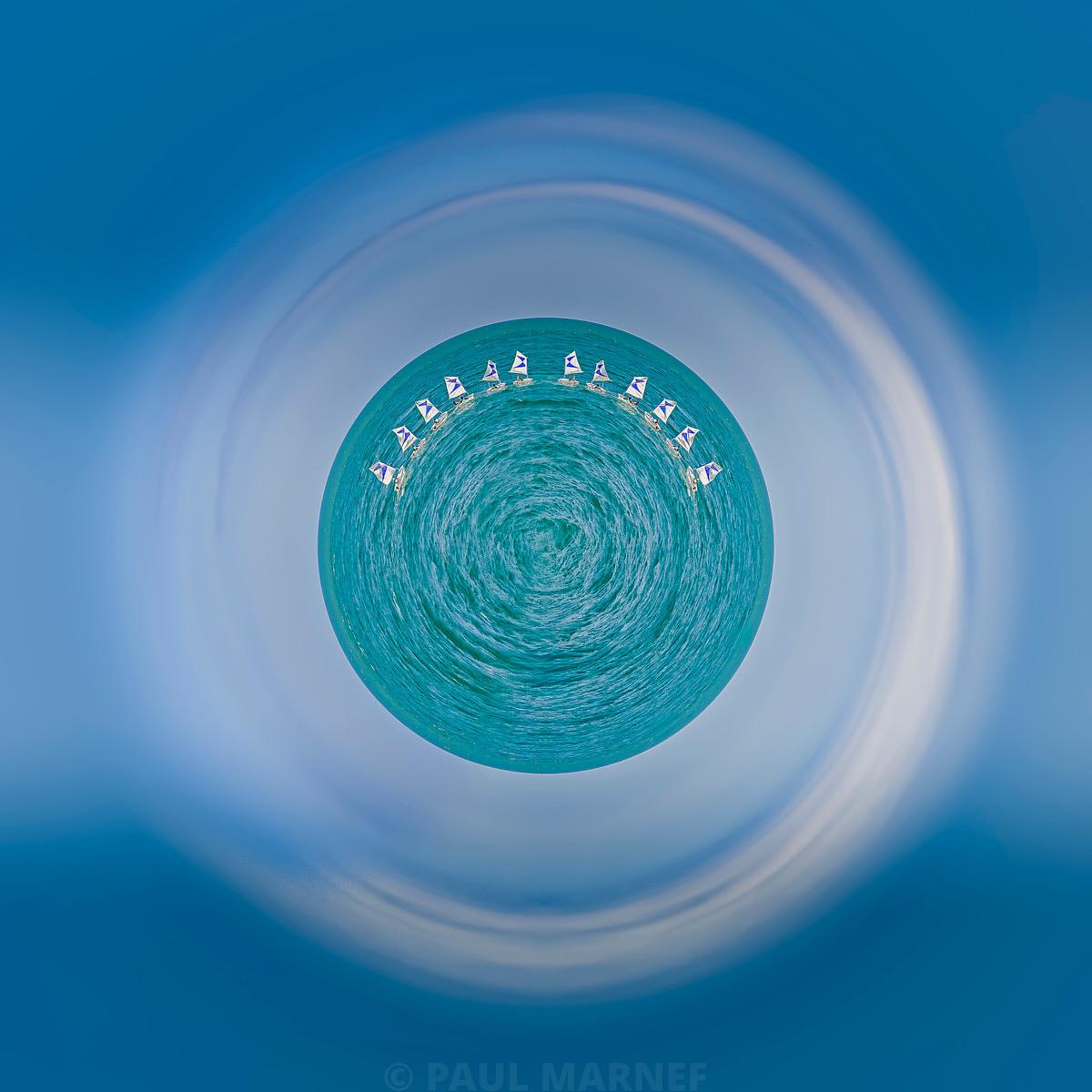 Une flottille de petits voiliers cerne un disque d'eau turquoise et tournoyante, entourée de larges flous circulaires dominés par le bleu ciel.