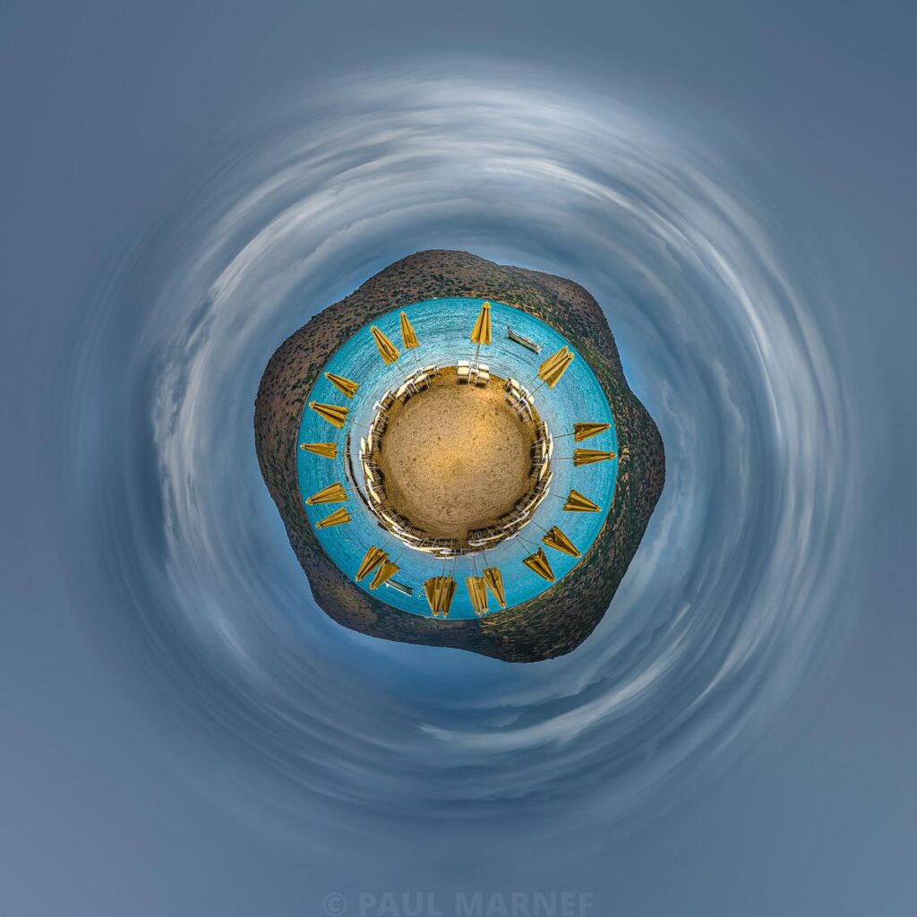 Seen through a fisheye lens, a turquoise beach lined with yellow parasols and framed by a cloudy sky forming white swirls.