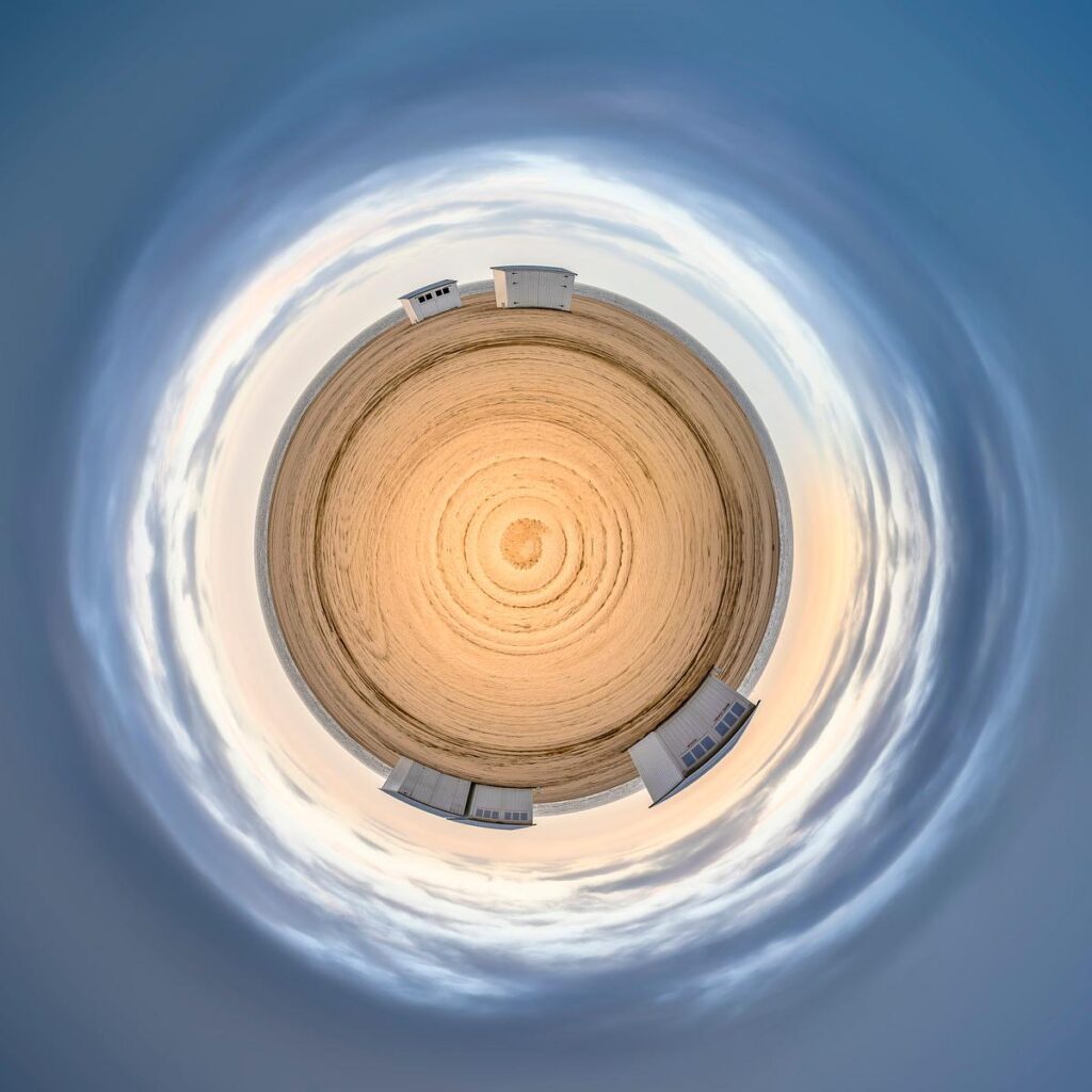 Plage de sable doré en cercle parfaite sous un ciel bleu vaporeux, entrecoupé de quelques petites bâtisses blanches.