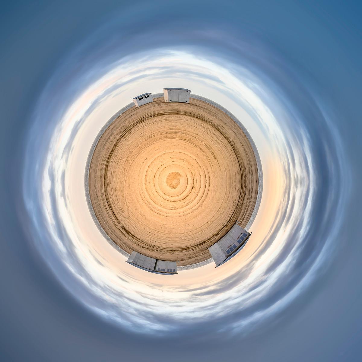 Plage de sable doré en cercle parfaite sous un ciel bleu vaporeux, entrecoupé de quelques petites bâtisses blanches.