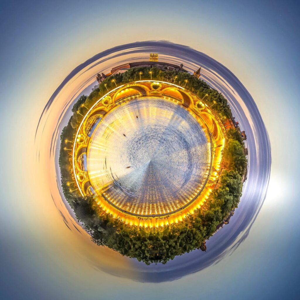 A panoramic night-time scene featuring arches bathed in golden light, lush green trees and a central square with silvery reflections.