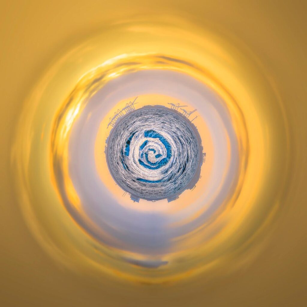 Sphère centrale composée d'un océan bleuté et de vagues blanches en spirale, surmontée de pales de turbines éoliennes, entourée d'un ciel incandescent aux nappes jaune-dorées.