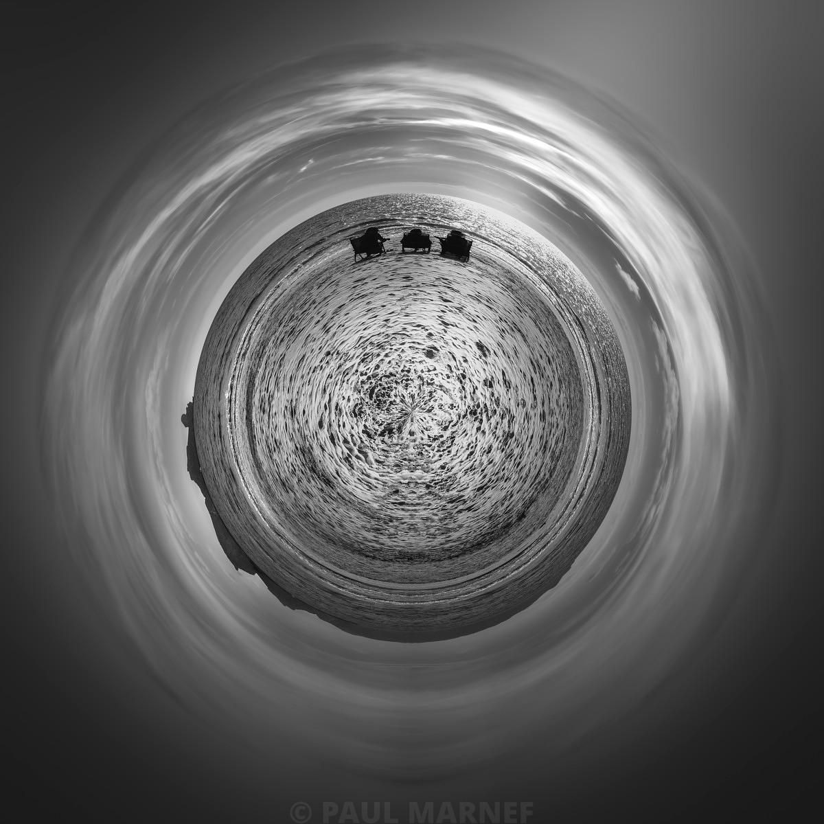 Vue en noir et blanc d'une plage sous un ciel aux nuages filés en spirale côtoyant trois silhouettes assises.