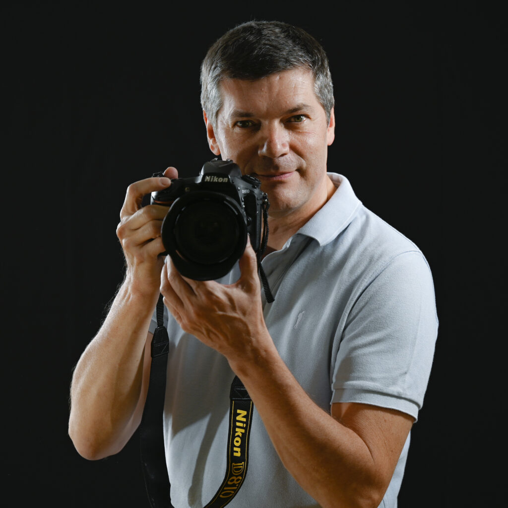 Paul Marnef holding a camera, wearing a blue T-shirt against a black background