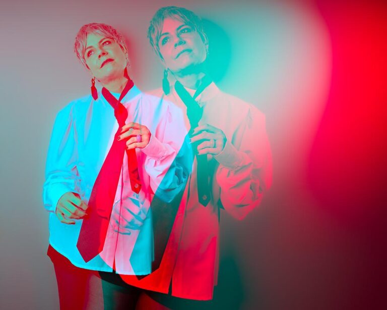 Ce portrait présente une femme dans une pose réfléchie, habillée d'une chemise blanche avec une cravate rouge lâche autour du cou. L'image utilise un effet visuel dynamique avec une double exposition ou un dédoublement des couleurs en rouge et bleu cyan, ce qui donne un effet de mouvement ou de réflexion, presque comme une illusion d'optique. Le contraste entre ces deux couleurs vibrantes crée une atmosphère à la fois artistique et rétro.  L'éclairage provient de plusieurs sources, générant des ombres colorées distinctes, renforçant l'effet psychédélique et moderne. La personne dans la photo adopte une expression douce et contemplative, regardant légèrement au loin, renforçant une ambiance introspective et un peu mystérieuse.  L'utilisation de ce style visuel avec les couleurs décalées et la duplication de l'image crée un sentiment de dualité ou de réflexion intérieure, suggérant une exploration de l'identité ou de l'émotion à travers la photographie.