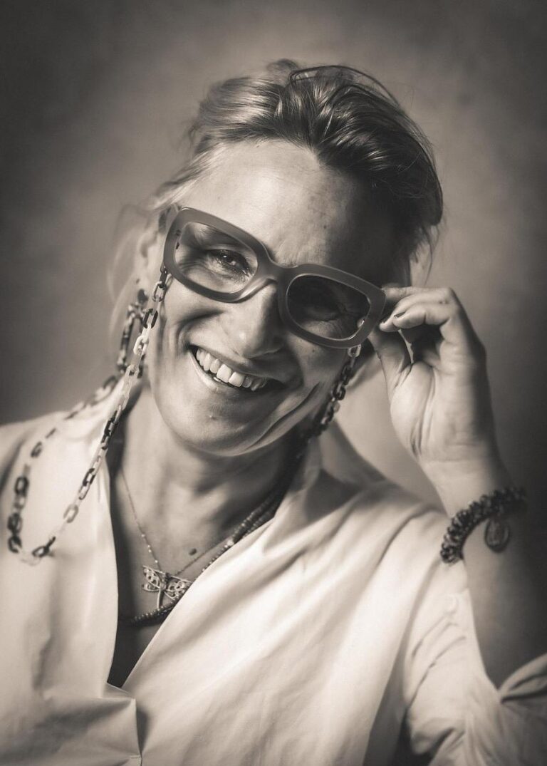 Portrait en noir et blanc d'une femme souriante ajustant ses lunettes, capturé par Paul Marnef, photographe portraitiste à Bruxelles, montrant un moment de spontanéité et de bonheur.