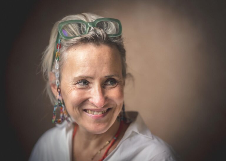 Portrait photo de Sarah Debeuckelaer par Paul Marnef, photographe professionnel. Le portrait photo montre une femme souriante. L'image est horizontale, en deux tons, presque noir et blanc, avec une retouche douce et professionnelle