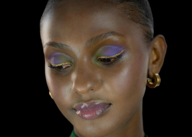 Portrait d'une femme avec un maquillage vibrant violet et or, capturé par Paul Marnef, photographe professionnel à Bruxelles. Le modèle regarde vers le bas avec un air doux et serein, sur un fond noir.