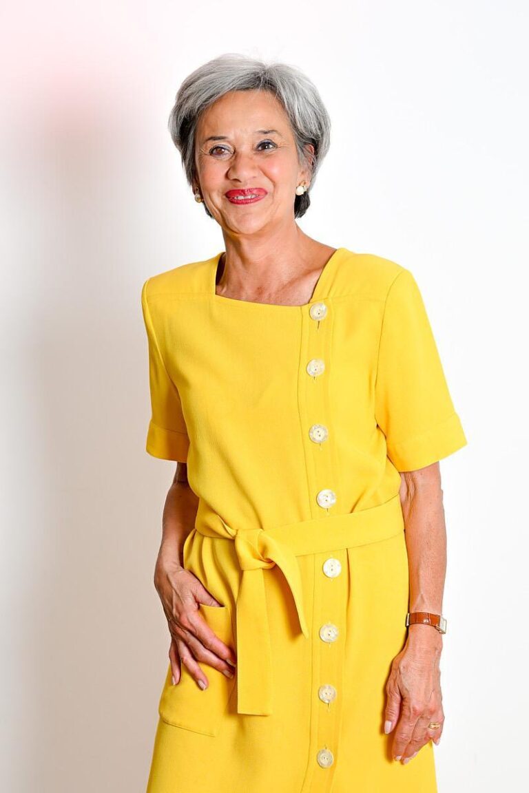 Portrait d'une femme élégante aux cheveux gris, vêtue d'une robe jaune vif, réalisé par Paul Marnef, photographe portraitiste professionnel à Bruxelles. Le fond clair et épuré met en valeur l'attitude confiante et souriante du modèle.