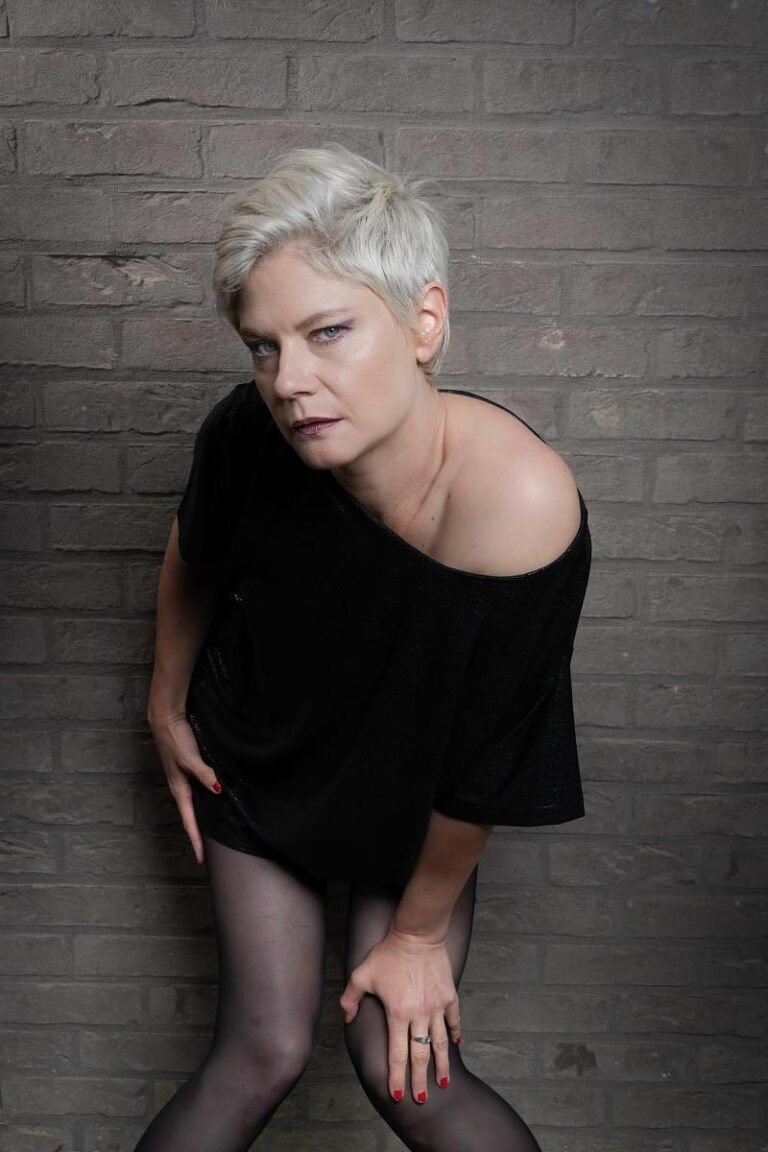 Portrait d'une femme dans un style rock intense, habillée en noir et adoptant une posture audacieuse devant un mur de briques, capturé par Paul Marnef, photographe professionnel à Bruxelles.